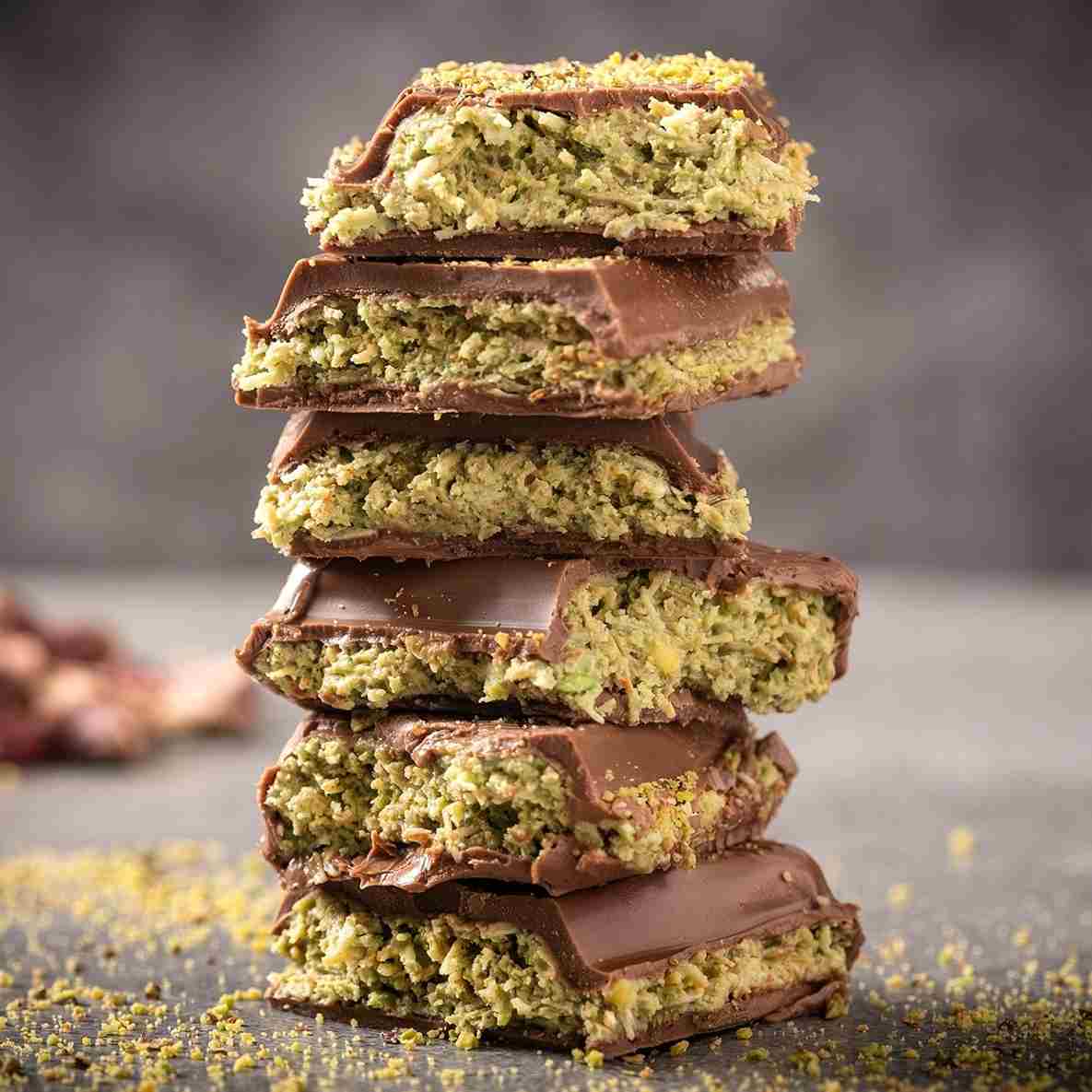 A stack of chocolate-covered bars with a green, nutty filling, likely made with pistachios, placed on a gray surface with some crumbs scattered around.